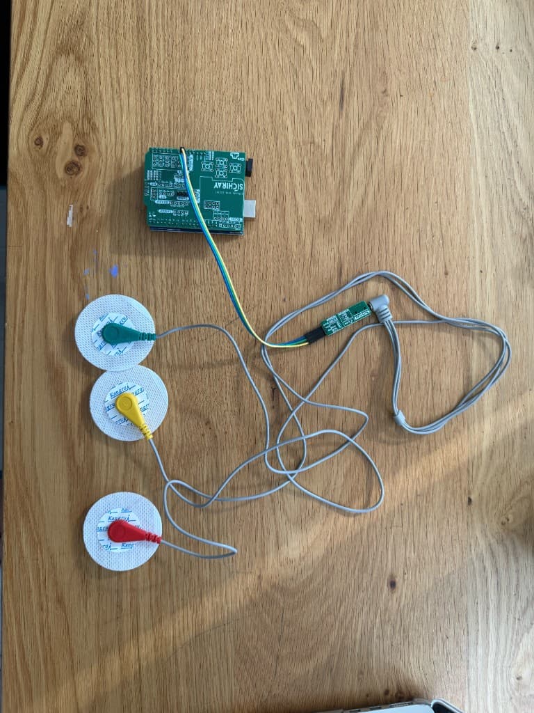 A green microcontroller board on a wooden table, wired to three round electrode pads with green, yellow, and red snaps, and a small green sensor board.