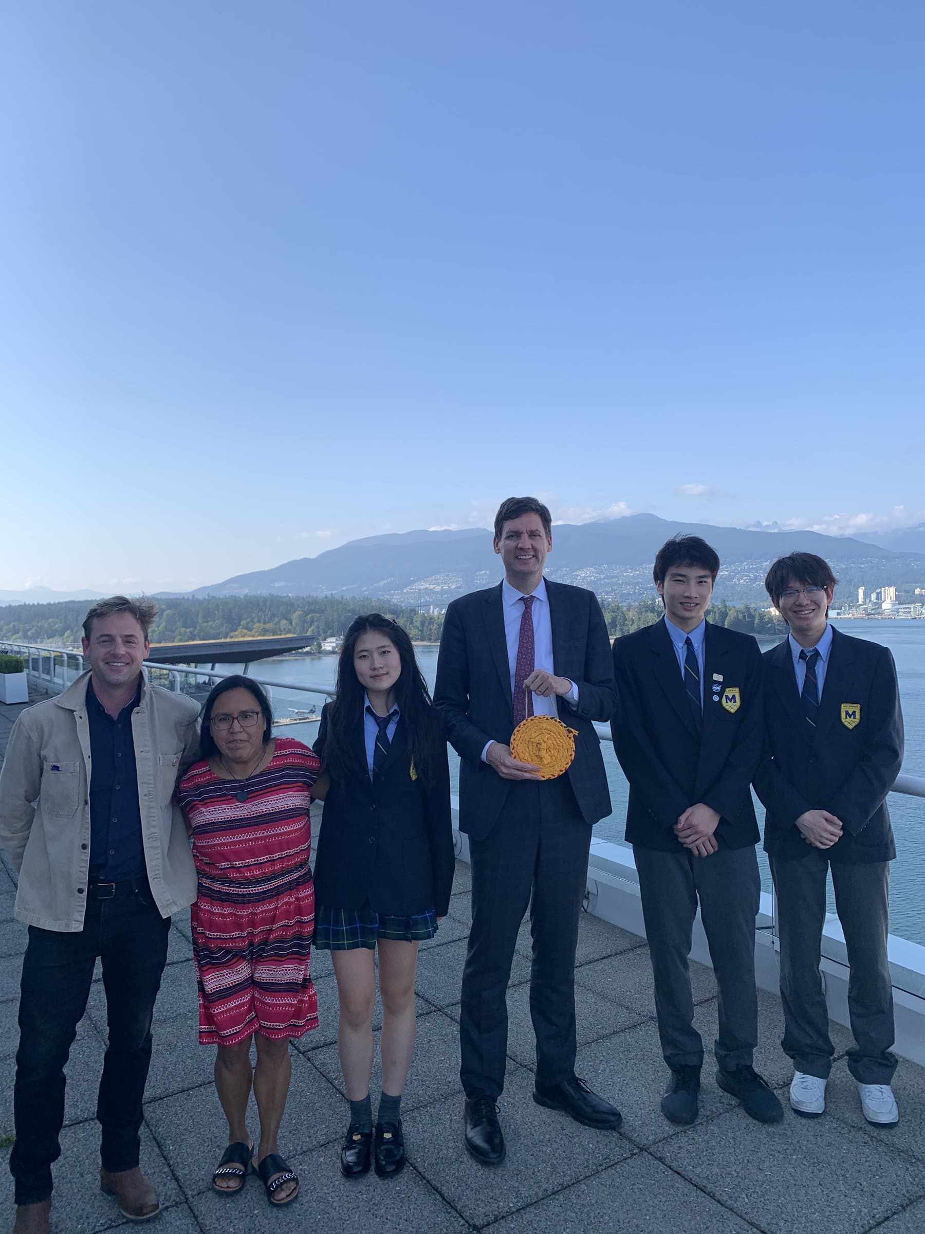 Six people on a paved terrace by the water, with mountains behind them. A man in a suit holds a round yellow object. Two students wear navy school blazers.