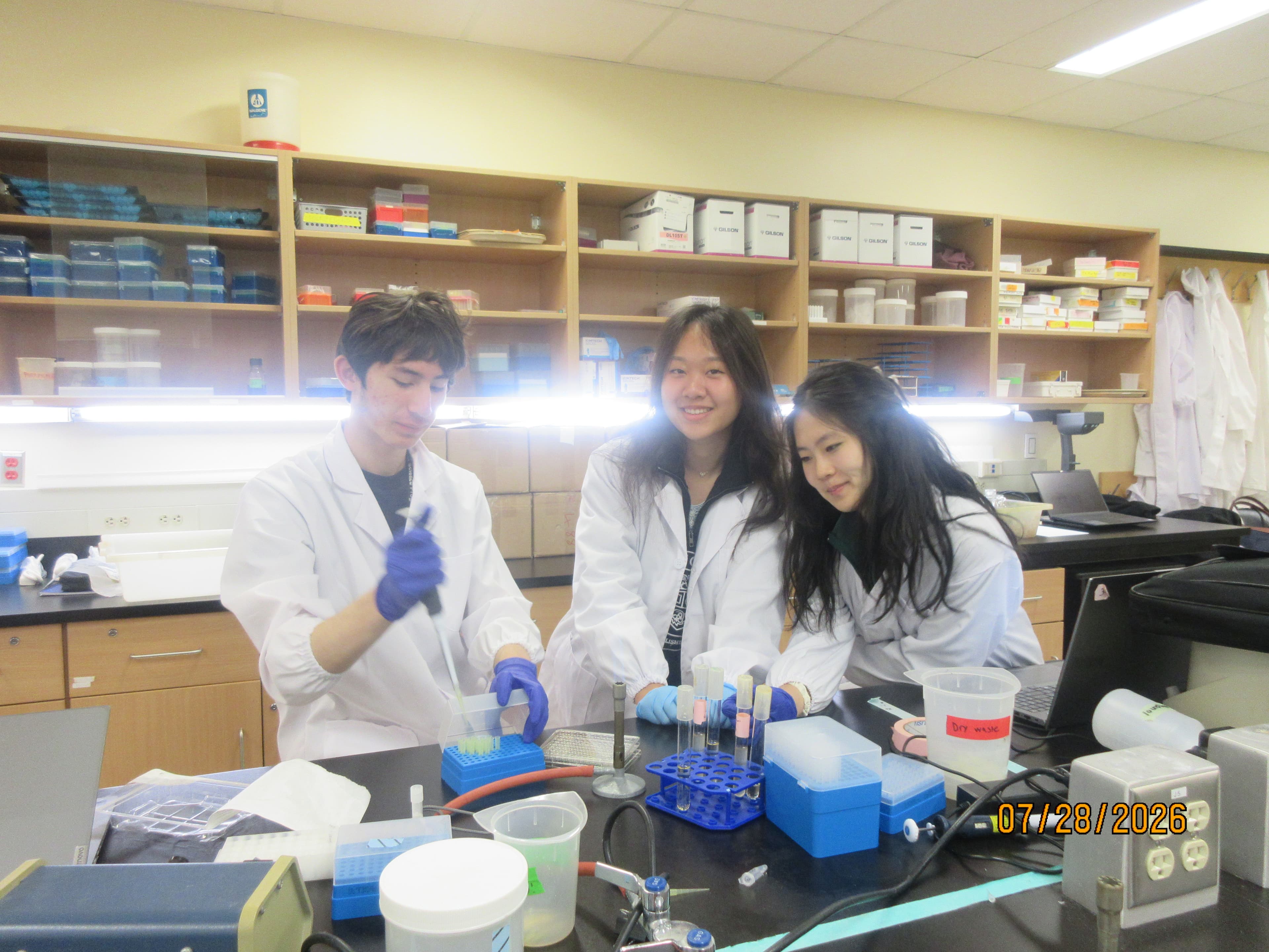 Three students in lab coats at a lab bench. One holds a pipette over a rack of tubes. The photo is stamped 07/28/2026.