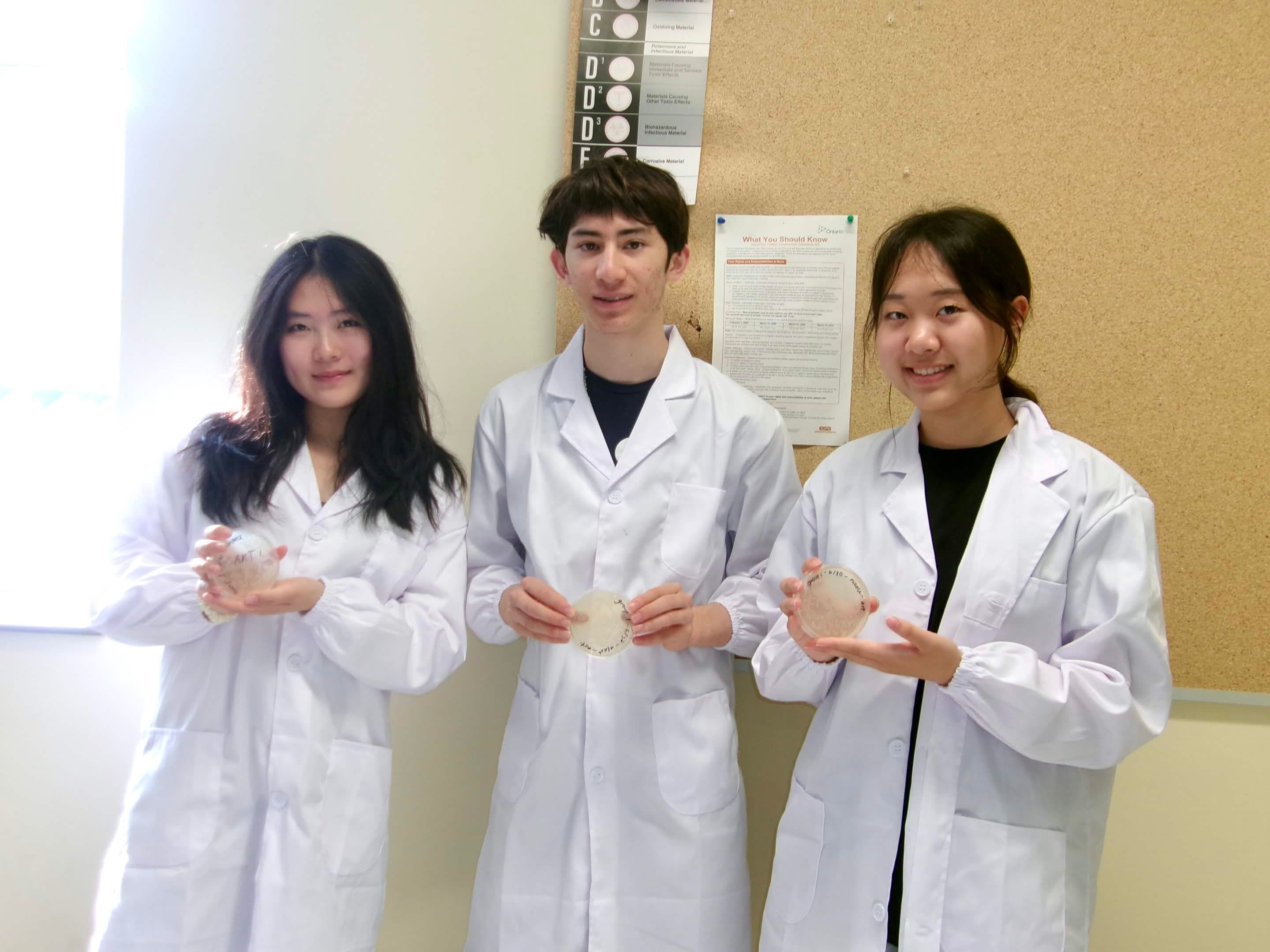 Three students in white lab coats holding petri dishes.
